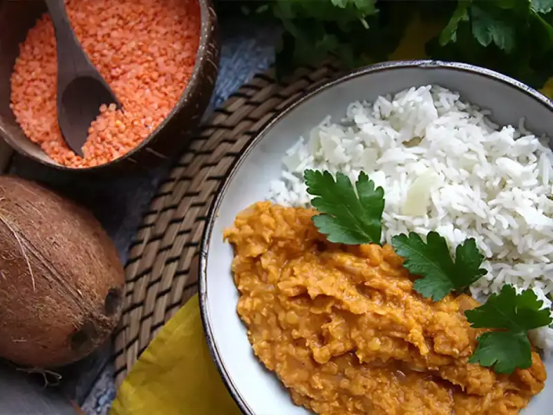 Piatto di riso basmati e curry di lenticchie rosse decorticate, ispirato a ricette esotiche. Accanto, una ciotola di lenticchie crude e un cocco.